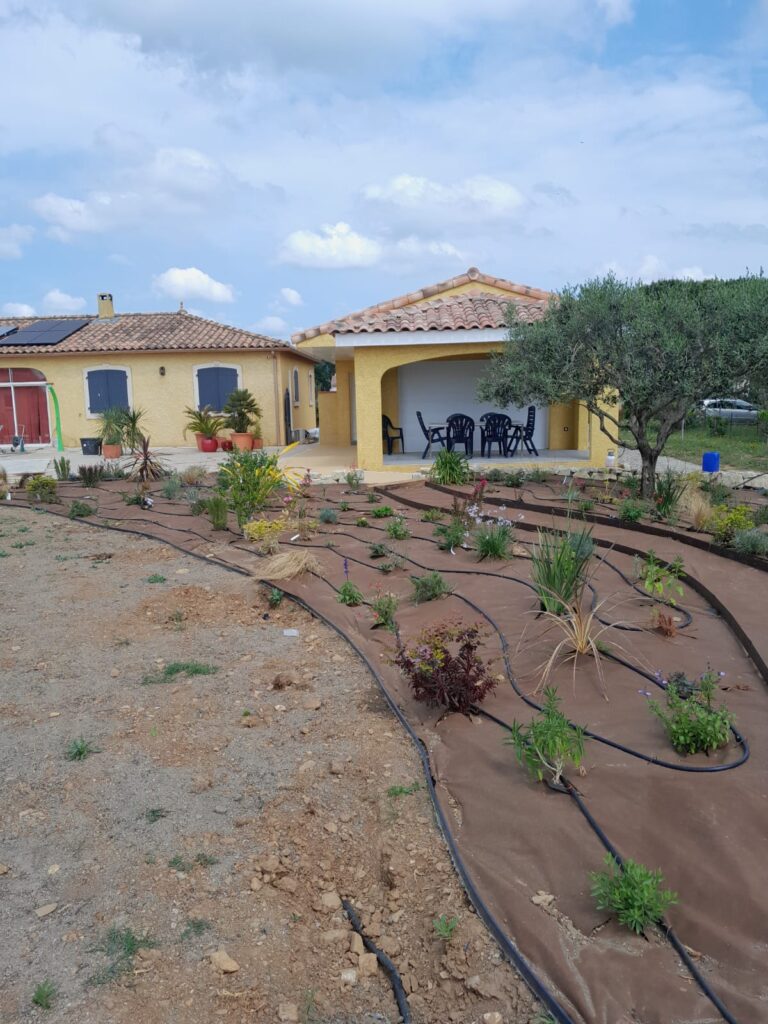 Aménagement de jardin méditerranéen avec système d'arrosage goutte-à-goutte et bâche de paillage devant une villa jaune.