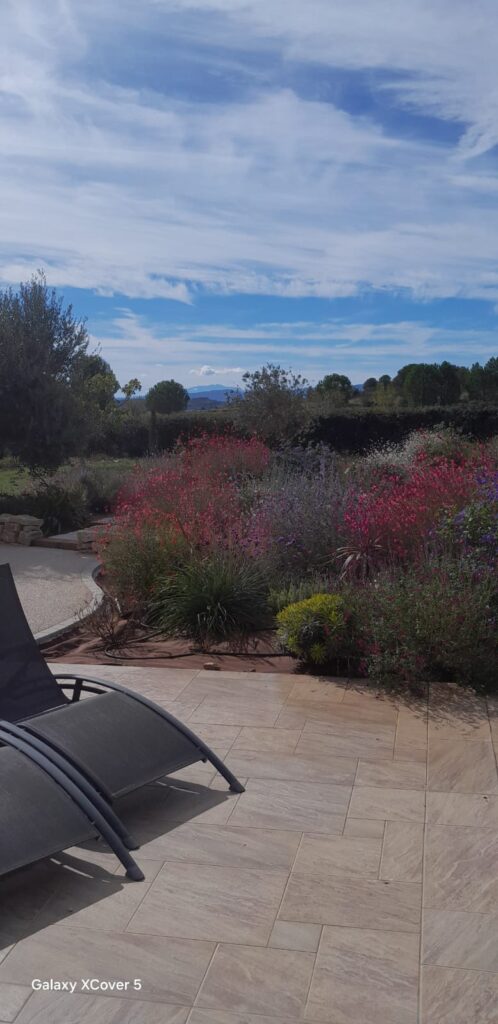 Terrasse en Pierre et Jardin Fleuri avec Vue Paysagère Terrasse en pierre avec transats devant un jardin fleuri rouge et violet sous un ciel bleu. Galaxy XCover 5.